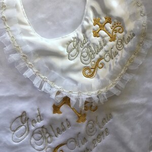Peut inclure: Bavoir de b&eacute;b&eacute; en satin blanc avec broderies et croix dor&eacute;es. Le texte brod&eacute; dit "God Bless Via Marie" et la date "April 8th, 2018". Une d&eacute;licate bordure en dentelle orne les bords.