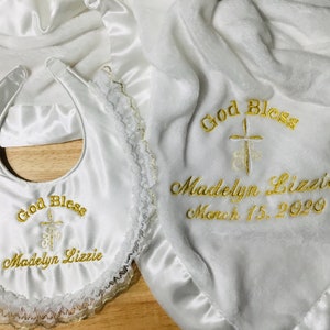 May include: White baby blanket, bib, and bonnet set. The blanket and bib are embroidered with "God Bless", a cross, and the name "Madelyn Lizzie" with the date March 15, 2020 in gold thread. The bib has a lace trim.