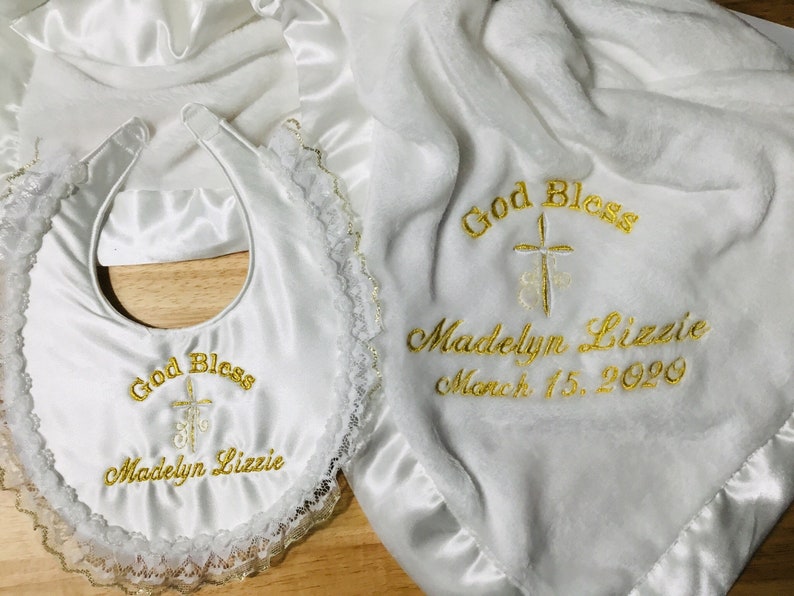 May include: White baby blanket, bib, and bonnet set. The blanket and bib are embroidered with "God Bless", a cross, and the name "Madelyn Lizzie" with the date March 15, 2020 in gold thread. The bib has a lace trim.