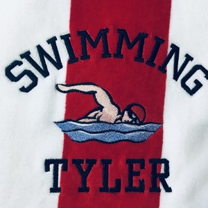 May include: White and red striped towel with the word "SWIMMING" arched across the top in navy blue embroidery. Below is an embroidered swimmer in the water, and the name "TYLER" is embroidered in navy blue at the bottom.