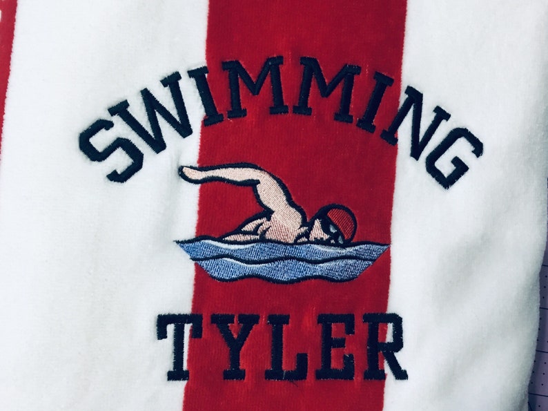 May include: White and red striped towel with the word "SWIMMING" arched across the top in navy blue embroidery. Below is an embroidered swimmer in the water, and the name "TYLER" is embroidered in navy blue at the bottom.