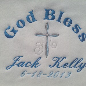 May include: White fabric embroidered with the words "God Bless" in blue script, arched across the top. A cross is centered below the words. The name "Jack Kelly" and the date "6-18-2013" are embroidered in blue script below the cross.