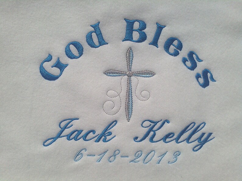May include: White fabric embroidered with the words "God Bless" in blue script, arched across the top. A cross is centered below the words. The name "Jack Kelly" and the date "6-18-2013" are embroidered in blue script below the cross.