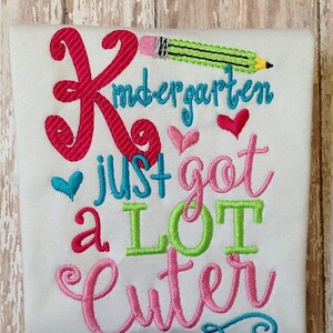 May include: White t-shirt with embroidered text and graphics. The design includes the word "Kindergarten" in teal, a large pink "K", a green pencil, and the phrase "just got a LOT cuter" in various colors with heart accents.