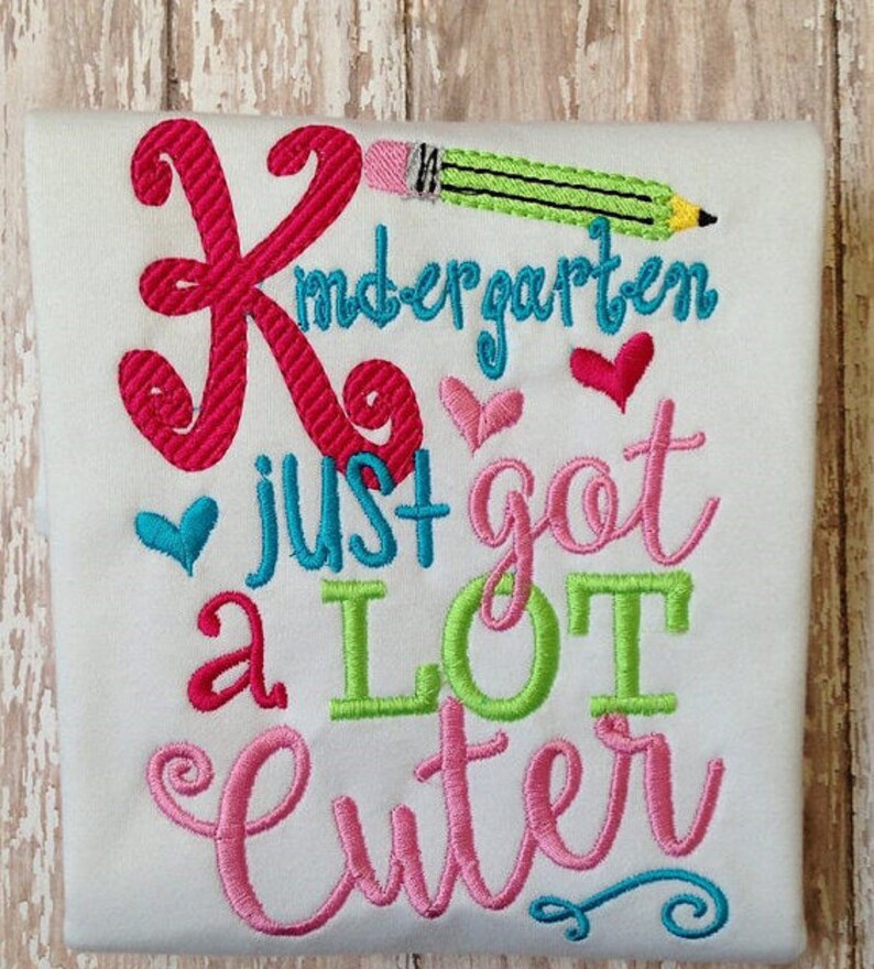 May include: White t-shirt with embroidered text and graphics. The design includes the word "Kindergarten" in teal, a large pink "K", a green pencil, and the phrase "just got a LOT cuter" in various colors with heart accents.