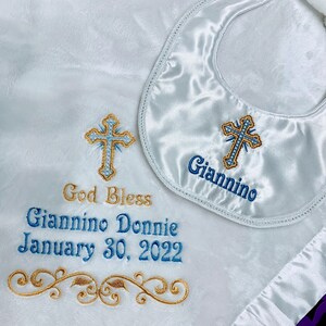May include: White satin baby blanket and bib set. The blanket features embroidered text that reads "God Bless Giannino Donnie January 30, 2022" and a gold and blue cross design. The bib has the name "Giannino" embroidered.