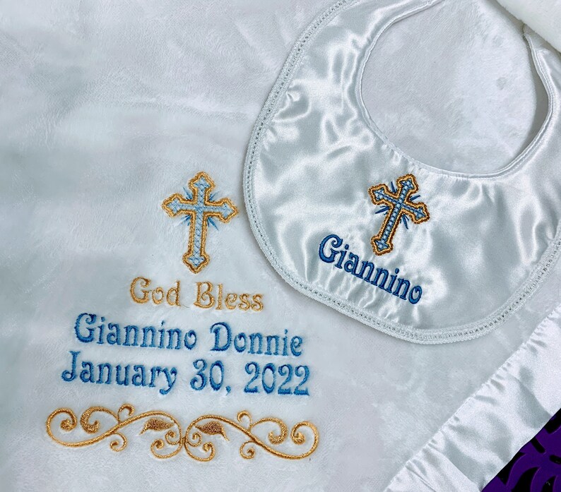 May include: White satin baby blanket and bib set. The blanket features embroidered text that reads "God Bless Giannino Donnie January 30, 2022" and a gold and blue cross design. The bib has the name "Giannino" embroidered.