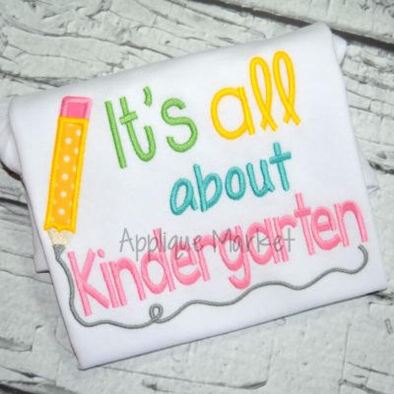 May include: White t-shirt with the embroidered text "It's all about Kindergarten" in green, yellow, blue, and pink. A yellow pencil with a pink eraser is on the left side. A gray line underlines the text.
