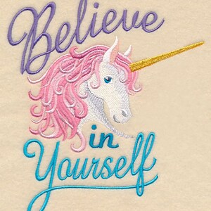 May include: Embroidered design featuring a unicorn with pink mane and a golden horn. The text "Believe in Yourself" is written above and below the unicorn in purple and teal script, respectively. The background is a light beige color.
