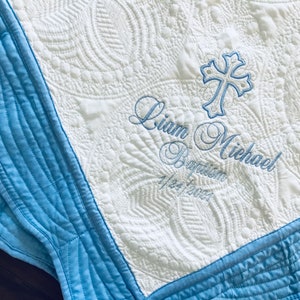 May include: White quilted baby blanket with blue trim and embroidered cross. The blanket features the name "Liam Michael", the word "Baptism", and the date "1/24/2023" embroidered in blue thread.