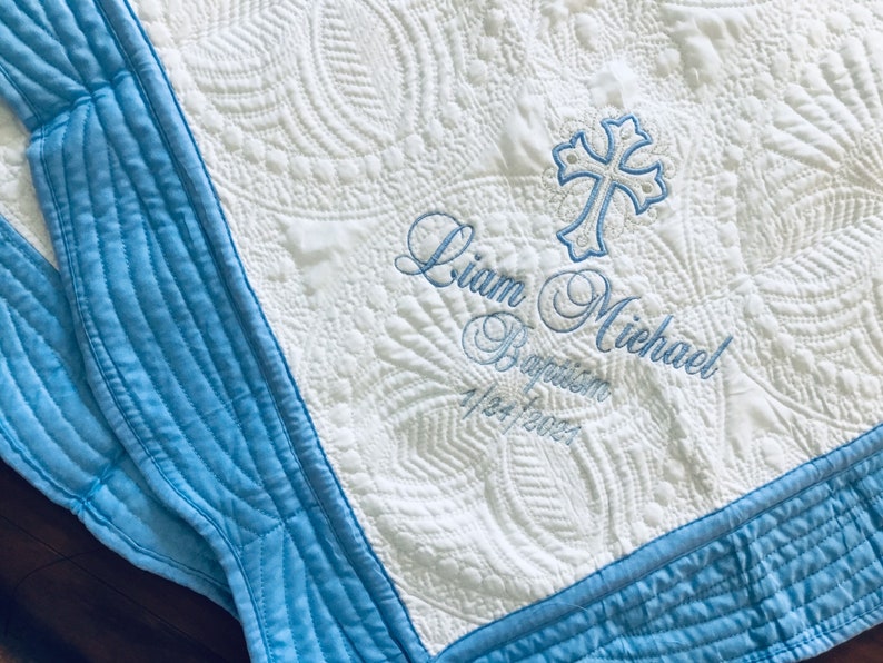 May include: White quilted baby blanket with blue trim and embroidered cross. The blanket features the name "Liam Michael", the word "Baptism", and the date "1/24/2023" embroidered in blue thread.