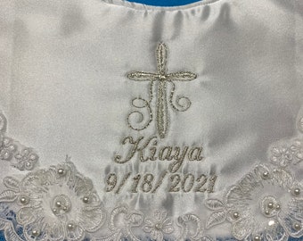 May include: White satin bib with a silver embroidered cross and the name "Kiaya" with the date "9/18/2021". The bib features a delicate lace trim with pearl accents.