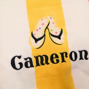 May include: White beach towel with a yellow stripe down the center. The towel features an embroidered pair of flip-flops with colorful polka dots and the name "Cameron" in black script. The towel is made of a soft, absorbent material.
