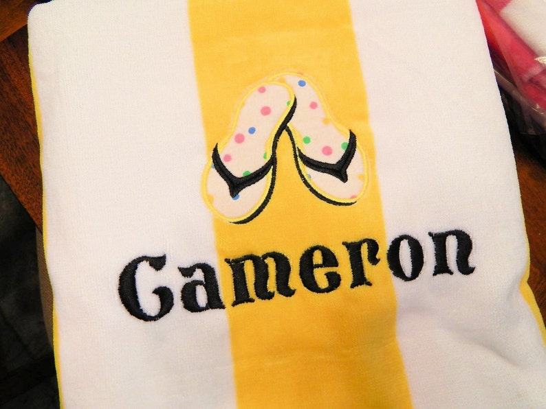 May include: White beach towel with a yellow stripe down the center. The towel features an embroidered pair of flip-flops with colorful polka dots and the name "Cameron" in black script. The towel is made of a soft, absorbent material.