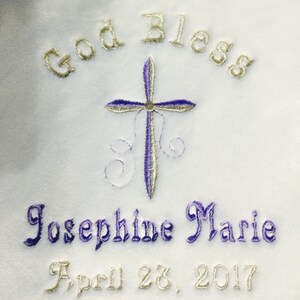 May include: White fabric embroidered with the words "God Bless" in gold thread above a cross with purple and silver detailing. Below the cross, the name "Josephine Marie" is embroidered in purple, and the date "April 28, 2017" is in gold.