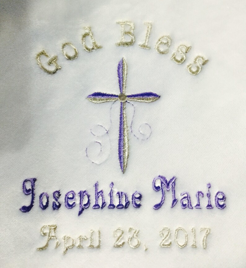 May include: White fabric embroidered with the words "God Bless" in gold thread above a cross with purple and silver detailing. Below the cross, the name "Josephine Marie" is embroidered in purple, and the date "April 28, 2017" is in gold.