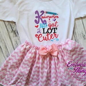 May include: White t-shirt and pink skirt set. The shirt has the text "Kindergarten just got a LOT Cuter" in colorful embroidery. The skirt is pink and white chevron patterned with a pink bow.