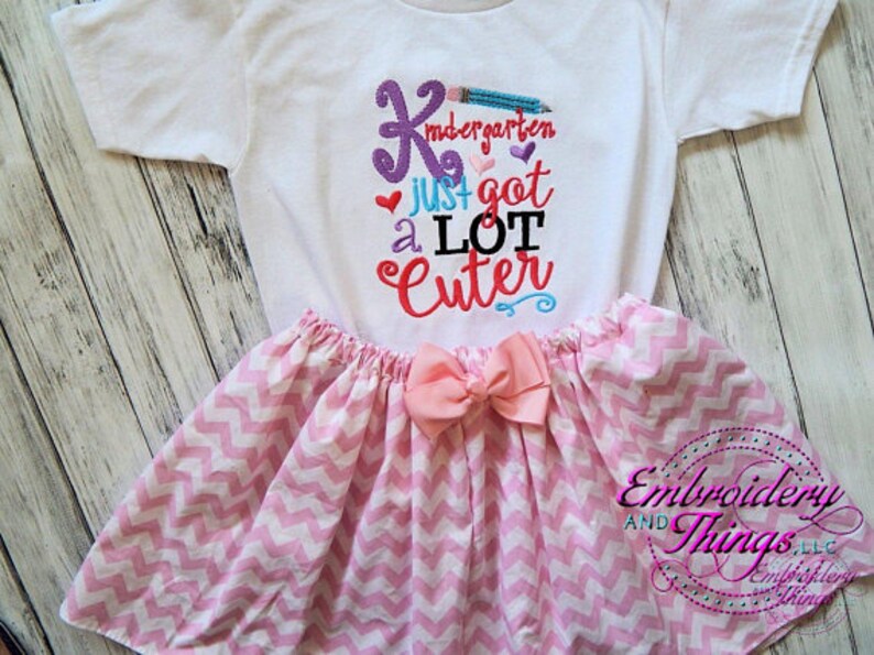 May include: White t-shirt and pink skirt set. The shirt has the text "Kindergarten just got a LOT Cuter" in colorful embroidery. The skirt is pink and white chevron patterned with a pink bow.