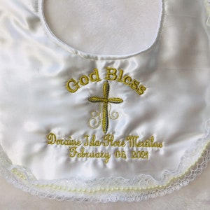 May include: White satin baby bib with embroidered gold text and cross design. The bib features the words "God Bless" and a name and date. The edges are trimmed with lace and small pearls.