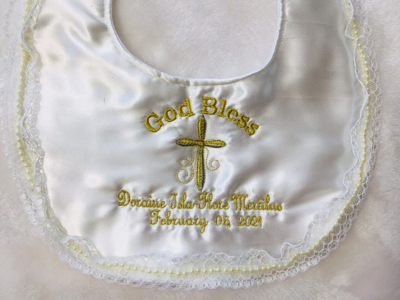 May include: White satin baby bib with embroidered gold text and cross design. The bib features the words "God Bless" and a name and date. The edges are trimmed with lace and small pearls.