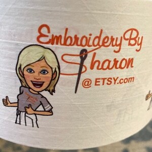 May include: White spool of thread with the text "Embroidery By Sharon @ Etsy.com" in orange. A cartoon image of a blonde woman giving a thumbs up is on the left side of the spool. A black needle is in the middle of the text.