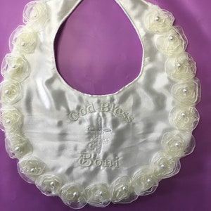 Peut inclure: Bavoir de b&eacute;b&eacute; en satin ivoire avec un bord festonn&eacute; de rosettes cr&egrave;me translucides, chacune centr&eacute;e d'une perle. Le bavoir porte la broderie "God Bless Boni" avec une croix. Fermeture velcro au cou.