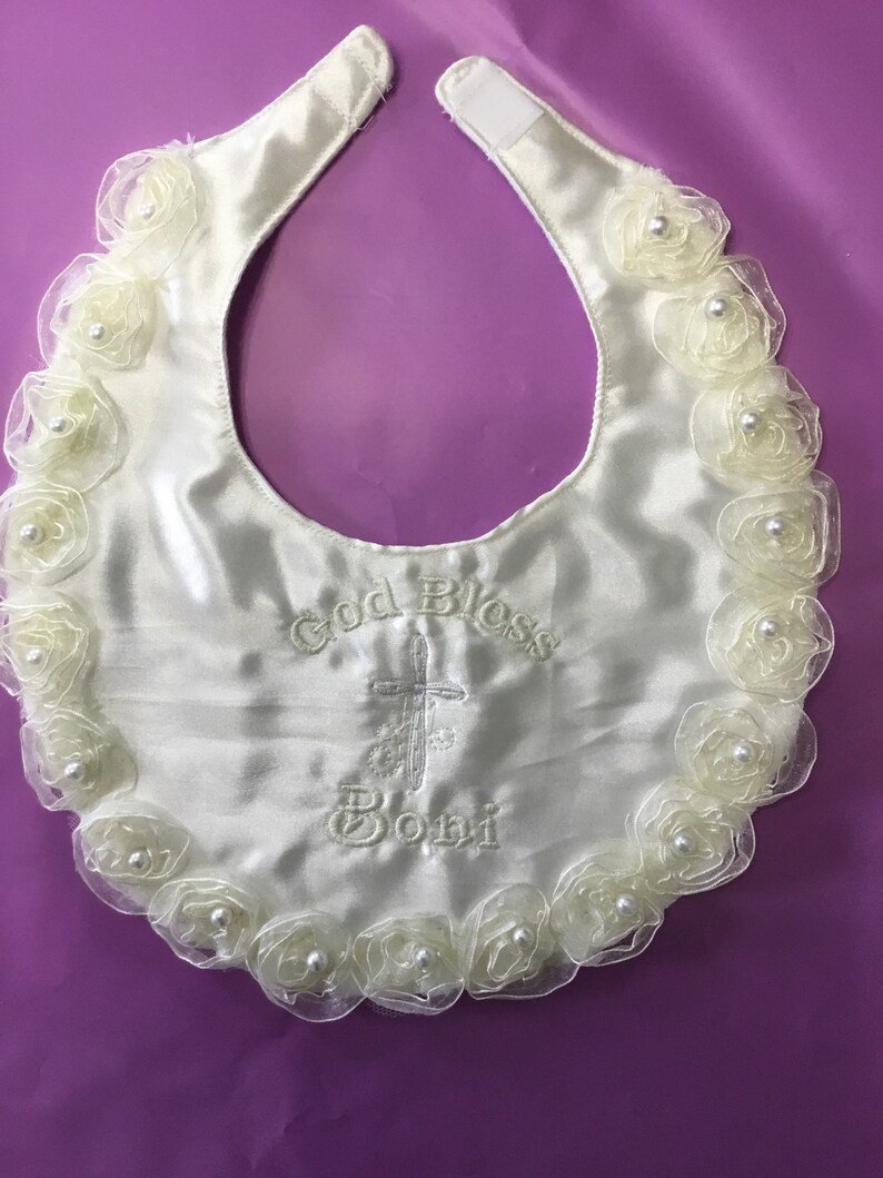 Peut inclure: Bavoir de b&eacute;b&eacute; en satin ivoire avec un bord festonn&eacute; de rosettes cr&egrave;me translucides, chacune centr&eacute;e d'une perle. Le bavoir porte la broderie "God Bless Boni" avec une croix. Fermeture velcro au cou.