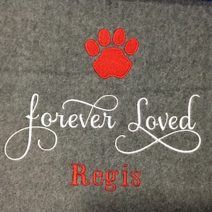 May include: A gray pet blanket with a red paw print and the embroidered text "forever Loved" in white script. Below, the name "Regis" is embroidered in red. The blanket is made of a soft, plush material.