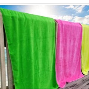 Puede incluir: Una fila de toallas de playa coloridas colgadas en una barandilla de madera. Las toallas son de tonos vibrantes de verde, rosa, amarillo y naranja. El fondo muestra un cielo azul con nubes y el mar. Las toallas son de material suave y absorbente.