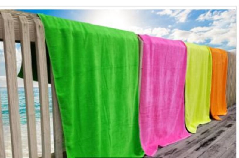 Puede incluir: Una fila de toallas de playa coloridas colgadas en una barandilla de madera. Las toallas son de tonos vibrantes de verde, rosa, amarillo y naranja. El fondo muestra un cielo azul con nubes y el mar. Las toallas son de material suave y absorbente.