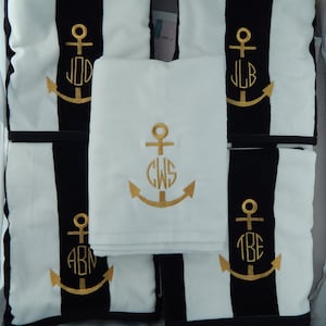 Puede incluir: Juego de cojines y una toalla con tem&aacute;tica n&aacute;utica. Los cojines presentan un dise&ntilde;o a rayas blancas y negras con anclas e iniciales bordadas en dorado. La toalla blanca del centro tambi&eacute;n tiene un ancla e iniciales doradas.