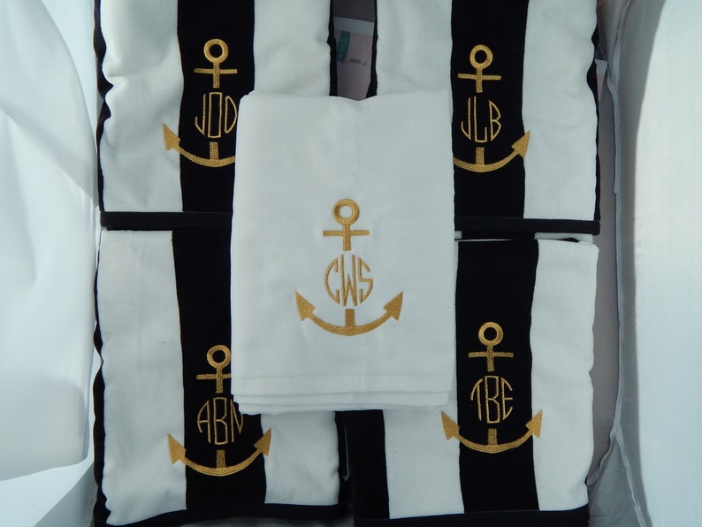 Puede incluir: Juego de cojines y una toalla con tem&aacute;tica n&aacute;utica. Los cojines presentan un dise&ntilde;o a rayas blancas y negras con anclas e iniciales bordadas en dorado. La toalla blanca del centro tambi&eacute;n tiene un ancla e iniciales doradas.