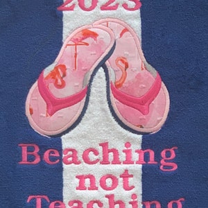 May include: A blue and white striped beach towel with embroidered pink text and flip-flops. The text reads "2023" above a pair of pink flip-flops with flamingo designs. Below the flip-flops, the text reads "Beaching not Teaching."