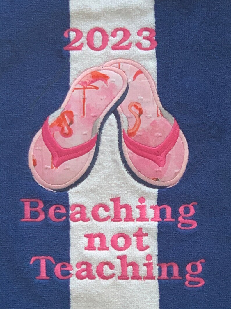 May include: A blue and white striped beach towel with embroidered pink text and flip-flops. The text reads "2023" above a pair of pink flip-flops with flamingo designs. Below the flip-flops, the text reads "Beaching not Teaching."