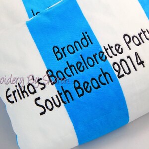 May include: A white and blue striped beach towel with black embroidered text. The text reads "Brandi Bachelorette Party South Beach 2014" and "Erika's". The towel is folded, showing the stripes and text.