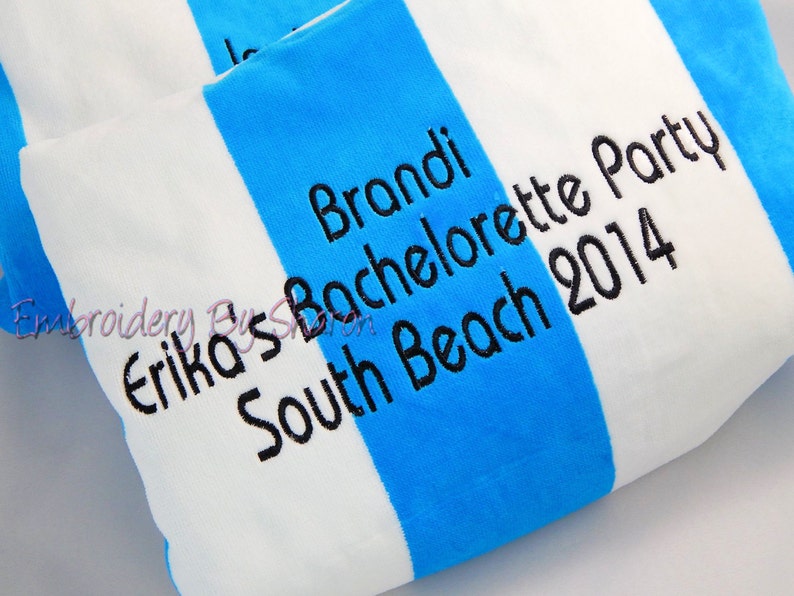 May include: A white and blue striped beach towel with black embroidered text. The text reads "Brandi Bachelorette Party South Beach 2014" and "Erika's". The towel is folded, showing the stripes and text.