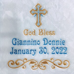 May include: Embroidered white fabric with a blue and gold cross at the top. The text "God Bless" is above the name "Giannino Donnie" and the date "January 30, 2022". A gold floral design is at the bottom. The fabric is framed in blue.