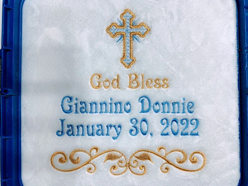 May include: Embroidered white fabric with a blue and gold cross at the top. The text "God Bless" is above the name "Giannino Donnie" and the date "January 30, 2022". A gold floral design is at the bottom. The fabric is framed in blue.
