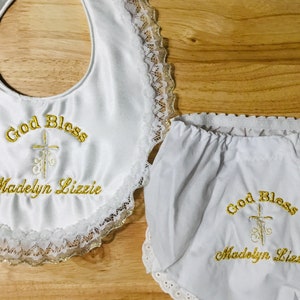 May include: White baby bib and diaper cover set with gold embroidered text and a cross design. The bib and diaper cover are trimmed with lace. The text on the bib and diaper cover reads "God Bless Madelyn Lizzie."