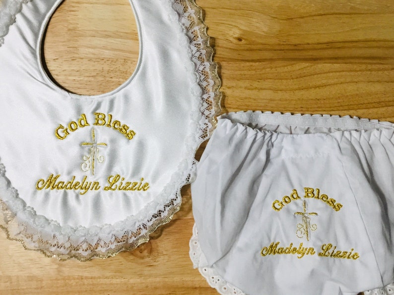 May include: White baby bib and diaper cover set with gold embroidered text and a cross design. The bib and diaper cover are trimmed with lace. The text on the bib and diaper cover reads "God Bless Madelyn Lizzie."
