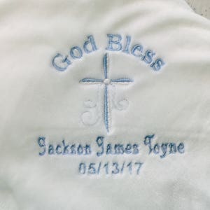 May include: White baby blanket with embroidered text and a cross design. The text reads "God Bless" above a light blue cross, with the name "Jackson James Toyne" and the date "05/13/17" embroidered below.