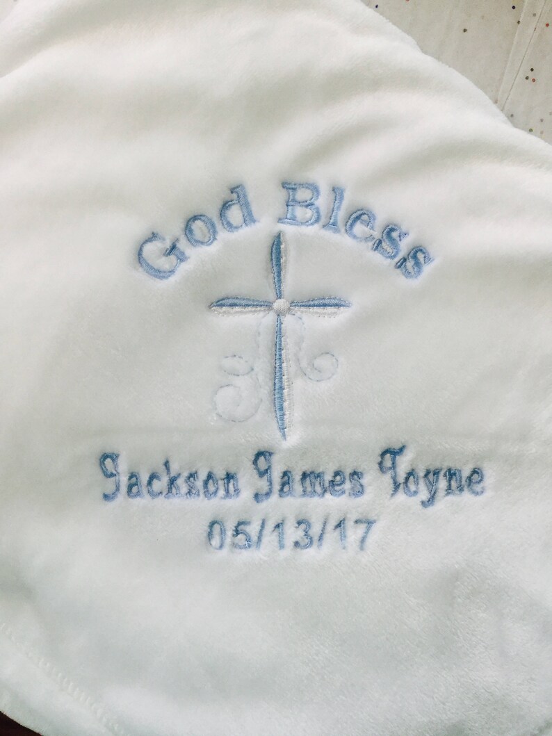 May include: White baby blanket with embroidered text and a cross design. The text reads "God Bless" above a light blue cross, with the name "Jackson James Toyne" and the date "05/13/17" embroidered below.
