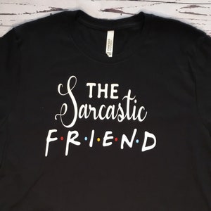 K&ouml;nnte beinhalten: Schwarzes T-Shirt mit dem wei&szlig;en Text "THE Sarcastic FRIEND", wobei das Wort "FRIEND" im Stil der Fernsehserie Friends gestaltet ist. Das T-Shirt besteht aus einem weichen Material und eignet sich f&uuml;r den Freizeitgebrauch.