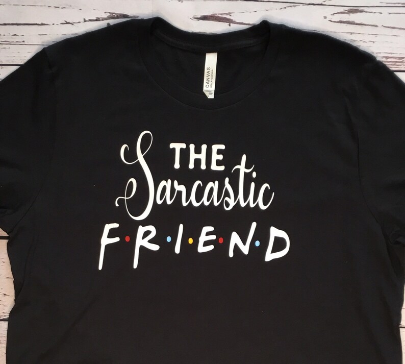 K&ouml;nnte beinhalten: Schwarzes T-Shirt mit dem wei&szlig;en Text "THE Sarcastic FRIEND", wobei das Wort "FRIEND" im Stil der Fernsehserie Friends gestaltet ist. Das T-Shirt besteht aus einem weichen Material und eignet sich f&uuml;r den Freizeitgebrauch.