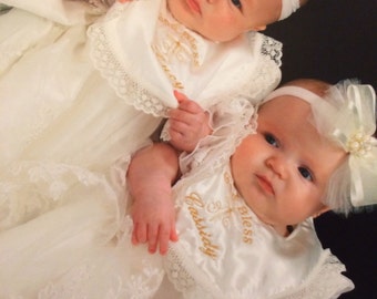May include: Two babies wearing white baptism gowns with lace trim and matching headbands. Each baby has a bib embroidered with a name and the word "Bless" in gold thread. The babies are positioned close together.