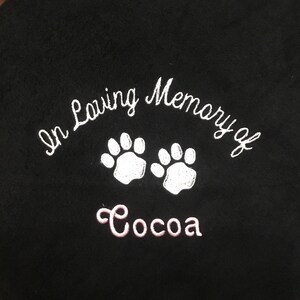 May include: Black blanket with white embroidered text that reads "In Loving Memory of" above two white paw prints. Below the paw prints is the name "Cocoa" embroidered in pink.