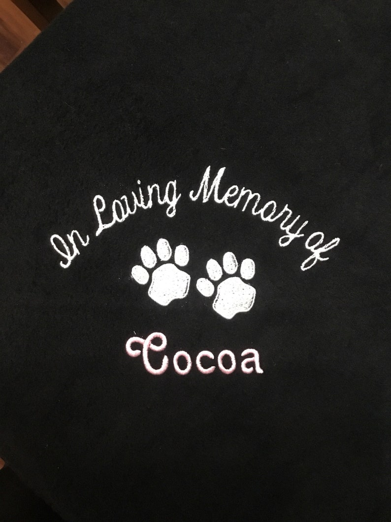 May include: Black blanket with white embroidered text that reads "In Loving Memory of" above two white paw prints. Below the paw prints is the name "Cocoa" embroidered in pink.