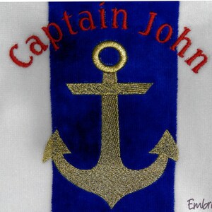 Puede incluir: Toalla blanca con una franja azul con un dise&ntilde;o de ancla dorada. Las palabras "Captain John" est&aacute;n bordadas en rojo encima del ancla. El dise&ntilde;o est&aacute; centrado en la toalla.