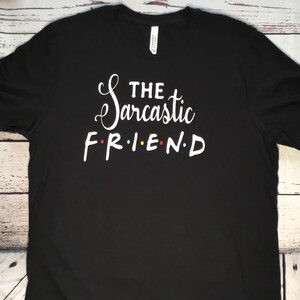 K&ouml;nnte beinhalten: Schwarzes T-Shirt mit dem wei&szlig;en Text "THE Sarcastic FRIEND". Das Wort "Sarcastic" ist kursiv geschrieben. Die Buchstaben in "FRIEND" sind rot, gelb, blau und rot.