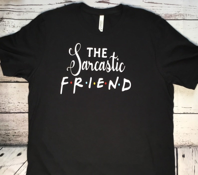 K&ouml;nnte beinhalten: Schwarzes T-Shirt mit dem wei&szlig;en Text "THE Sarcastic FRIEND". Das Wort "Sarcastic" ist kursiv geschrieben. Die Buchstaben in "FRIEND" sind rot, gelb, blau und rot.